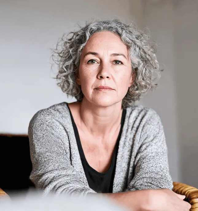 Een vrouw van middelbare leeftijd met grijs krullend haar kijkt rustig in de camera.